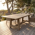 Full view of the Coastal Teak Dining Table outdoors on a sunlit patio, emphasizing length, wood grain, and rustic features against a coastal landscape.